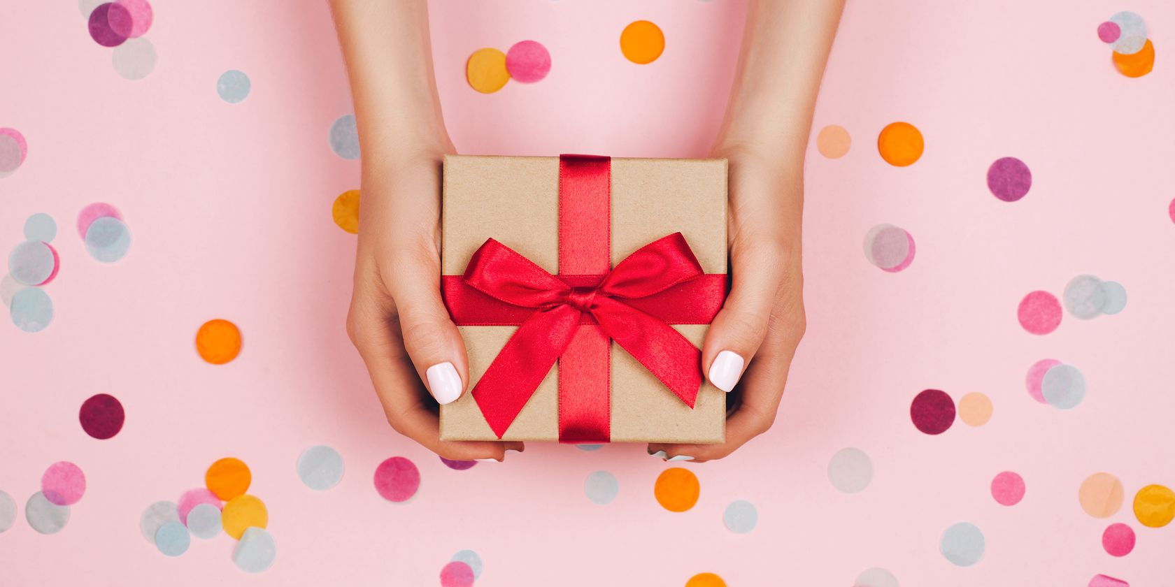 Woman's hands holding present box with red bow on pastel pink background with multicolored confetti